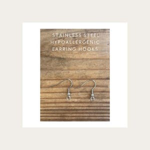 Peut inclure: Une paire de crochets de boucles d'oreilles en acier inoxydable de couleur argent&eacute;e avec un design en fil courb&eacute; et une petite boucle &agrave; la base. Le texte STAINLESS STEEL HYPOALLERGENIC EARRING HOOKS est imprim&eacute; en lettres capitales blanches.