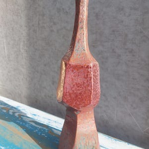 Puede incluir: Un jarrón de cerámica alto y artesanal con un diseño geométrico único. El jarrón presenta un color marrón rojizo con superficies texturizadas y detalles dorados. Su cuello alargado y cuerpo facetado le dan un atractivo moderno y artístico. El jarrón se exhibe sobre una superficie de madera azul y blanca.