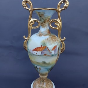 Puede incluir: Un jarrón decorativo con una escena de paisaje. El jarrón presenta un fondo azul claro con una escena pastoral pintada de una granja con un tejado rojo. El jarrón tiene asas doradas y un borde dorado. La base es blanca con un patrón marrón y dorado.