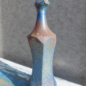 Puede incluir: Una botella decorativa de cerámica alta con un diseño geométrico. La botella presenta una combinación de colores azul y marrón con una superficie texturizada. La forma es alargada con una parte superior puntiaguda y una base ancha.