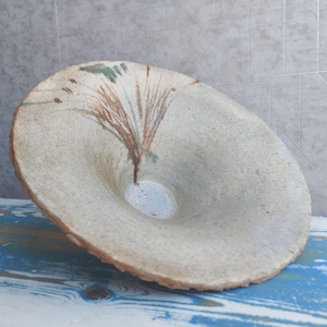 Puede incluir: Un cuenco de cerámica hecho a mano con un diseño ancho y poco profundo. El cuenco es de color beige claro con acentos abstractos pintados en marrón y verde. Se coloca sobre una superficie de madera azul y blanca. Diámetro: aprox. 30 cm.