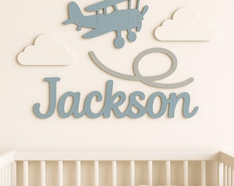 Puede incluir: Un conjunto de decoración mural para guardería con un avión azul, dos nubes blancas y el nombre "Jackson" en escritura azul. La decoración está montada sobre una cuna blanca.