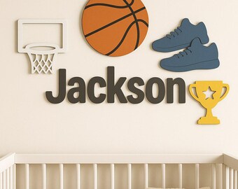 Könnte beinhalten: Wanddekoration mit einem Basketball, blauen Turnschuhen, einer gelben Trophäe und dem Namen "Jackson" in grauen Buchstaben. Der Basketball ist orange mit schwarzen Linien. Die Dekoration befindet sich über einem weißen Kinderbett.