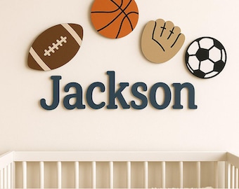 Könnte beinhalten: Wanddekoration mit einem Football, Basketball, Baseballhandschuh und Fußball über dem Namen "Jackson" in Marineblau. Die Sportdekorationen sind an einer weißen Wand angebracht.