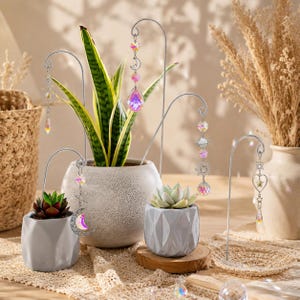 May include: A collection of crystal suncatchers suspended from metal stands, displayed with potted succulents and a snake plant. The crystals shimmer with multiple colours. The pots are grey and white, set against a neutral backdrop.