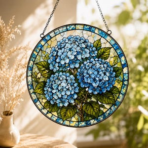 May include: A round stained glass window featuring a cluster of blue hydrangea flowers with green leaves. The flowers are in the center, surrounded by a mosaic-style border of blue, green, and yellow glass. A black chain is attached for hanging.
