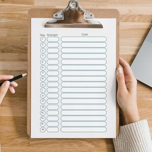 May include: A clipboard with a white paper checklist. The checklist has numbered rows and columns labeled "Day", "Strength", and "Goal". A hand holds a pencil, and another hand holds the clipboard. A laptop is visible in the background.
