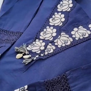 May include: A navy blue garment with white floral embroidery. The embroidery is featured on the neckline, sleeves, and other areas of the garment. The garment has a tassel detail and a lace trim.