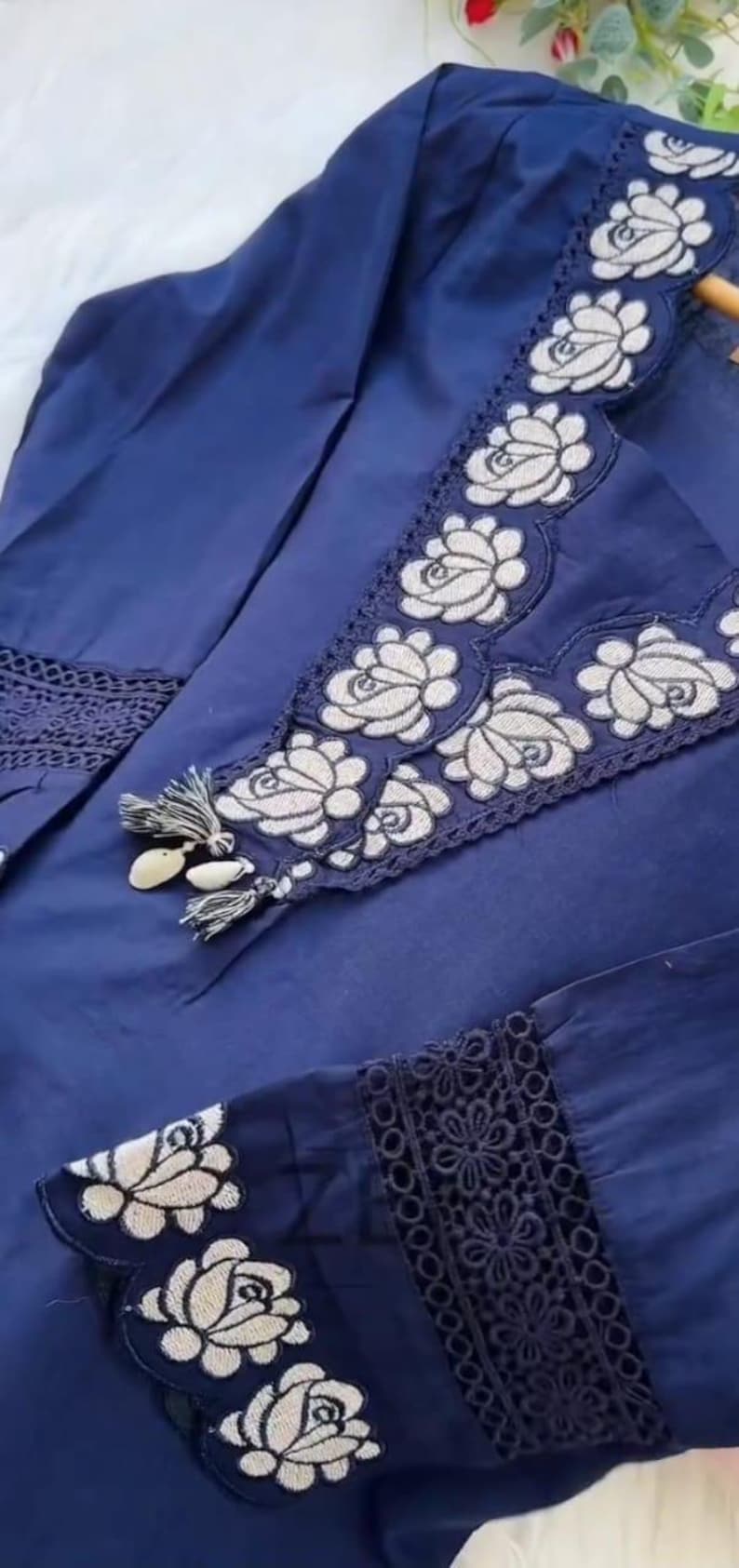 May include: A navy blue garment with white floral embroidery. The embroidery is featured on the neckline, sleeves, and other areas of the garment. The garment has a tassel detail and a lace trim.