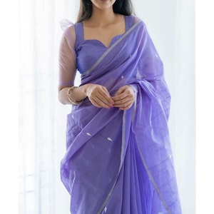 Puede incluir: Un sari lavanda con una blusa a juego. El sari presenta un patrón sutil y un borde plateado. La blusa tiene un escote corazón y mangas transparentes. El conjunto está hecho de una tela ligera.