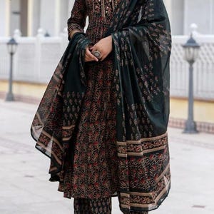 May include: A dark green and black printed salwar kameez set. The ensemble includes a long-sleeved tunic, matching pants, and a flowing scarf. The fabric features a floral pattern in red and gold accents. The outfit is paired with sandals.
