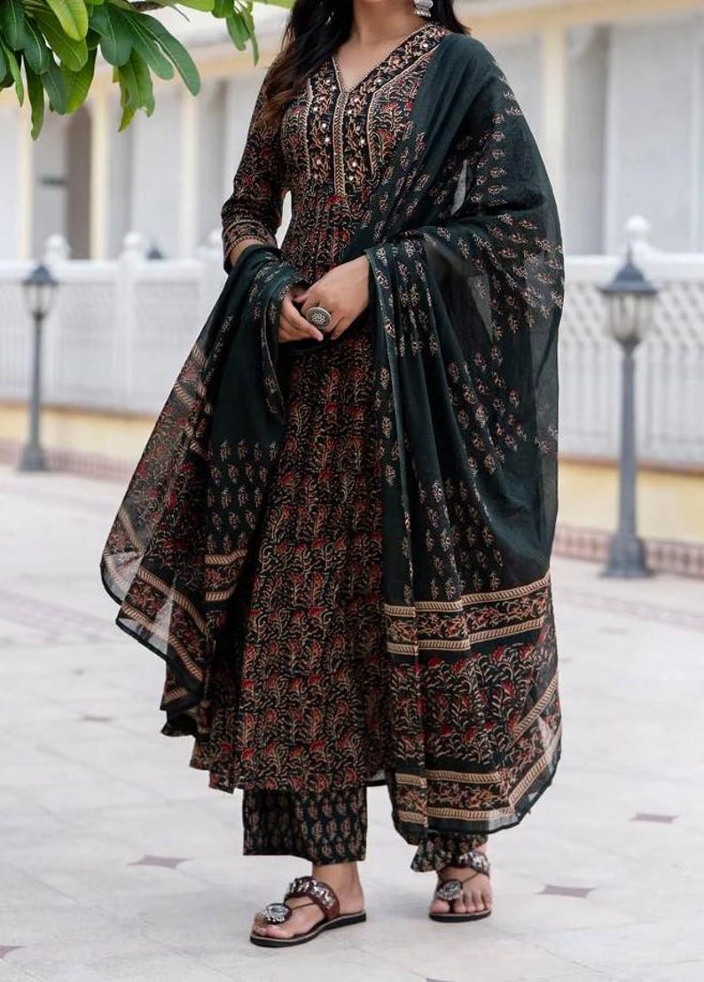 May include: A dark green and black printed salwar kameez set. The ensemble includes a long-sleeved tunic, matching pants, and a flowing scarf. The fabric features a floral pattern in red and gold accents. The outfit is paired with sandals.