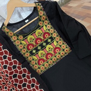 Puede incluir: T&uacute;nica negra con cuello bordado colorido. El bordado presenta motivos florales rojos, amarillos y verdes con peque&ntilde;os espejos. Una bufanda estampada en rojo, negro y blanco est&aacute; drapeada sobre el hombro.