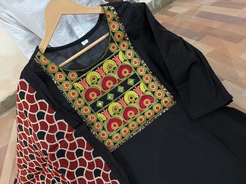 Puede incluir: T&uacute;nica negra con cuello bordado colorido. El bordado presenta motivos florales rojos, amarillos y verdes con peque&ntilde;os espejos. Una bufanda estampada en rojo, negro y blanco est&aacute; drapeada sobre el hombro.