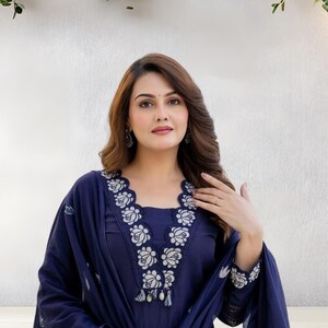 May include: A navy blue embroidered salwar kameez. The neckline and sleeves are adorned with white floral embroidery. The outfit includes a matching dupatta. The woman is wearing earrings and a ring.