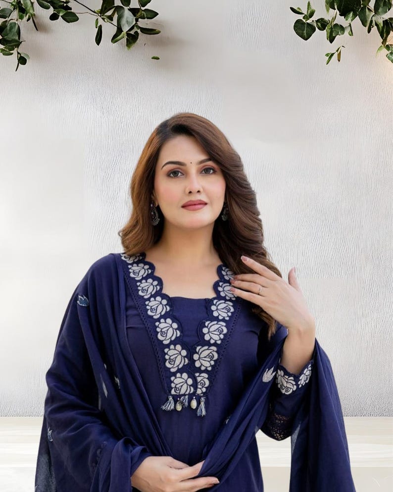 May include: A navy blue embroidered salwar kameez. The neckline and sleeves are adorned with white floral embroidery. The outfit includes a matching dupatta. The woman is wearing earrings and a ring.