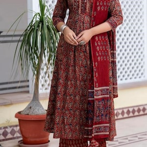 May include: A three-piece maroon Indian suit with a floral pattern. The set includes a long-sleeved tunic, matching pants, and a scarf. The outfit is adorned with decorative details around the neckline and cuffs. The model is wearing sandals.