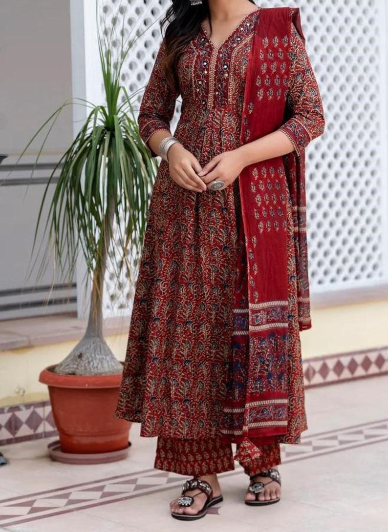 May include: A three-piece maroon Indian suit with a floral pattern. The set includes a long-sleeved tunic, matching pants, and a scarf. The outfit is adorned with decorative details around the neckline and cuffs. The model is wearing sandals.