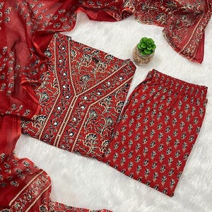 May include: A red Indian suit set featuring a tunic top, pants, and a sheer scarf. The set is adorned with a floral pattern in black and cream. The tunic has a V-neckline with mirror accents. The pants have a matching floral print.