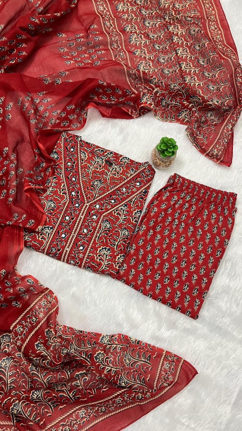 May include: A red Indian suit set featuring a tunic top, pants, and a sheer scarf. The set is adorned with a floral pattern in black and cream. The tunic has a V-neckline with mirror accents. The pants have a matching floral print.