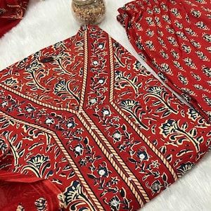 May include: A red floral print outfit, including a top, pants, and scarf. The top features a V-neck design with cream-colored trim and a repeating floral pattern in navy and white. The pants and scarf have a similar repeating pattern. The fabric appears soft.