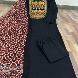 Puede incluir: Un conjunto de kurta negro con un pantal&oacute;n a juego y una bufanda estampada. El kurta presenta un dise&ntilde;o bordado colorido en el escote. La bufanda tiene un patr&oacute;n geom&eacute;trico rojo, negro y blanco.