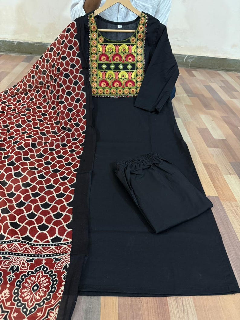 Puede incluir: Un conjunto de kurta negro con un pantal&oacute;n a juego y una bufanda estampada. El kurta presenta un dise&ntilde;o bordado colorido en el escote. La bufanda tiene un patr&oacute;n geom&eacute;trico rojo, negro y blanco.