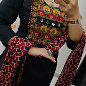 Puede incluir: T&uacute;nica negra de manga larga con bordados coloridos alrededor del escote. Los bordados incluyen detalles en rojo, amarillo y verde. Una bufanda a juego con un patr&oacute;n rojo y negro est&aacute; drapeada sobre los brazos.