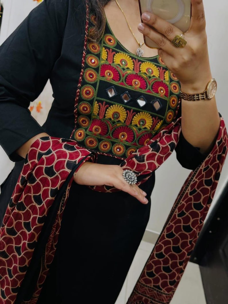 Puede incluir: T&uacute;nica negra de manga larga con bordados coloridos alrededor del escote. Los bordados incluyen detalles en rojo, amarillo y verde. Una bufanda a juego con un patr&oacute;n rojo y negro est&aacute; drapeada sobre los brazos.
