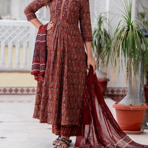 May include: A red floral print salwar kameez set. The long-sleeved top features a V-neck with decorative buttons and embroidery. The set includes matching pants and a sheer scarf. The outfit is worn with sandals.