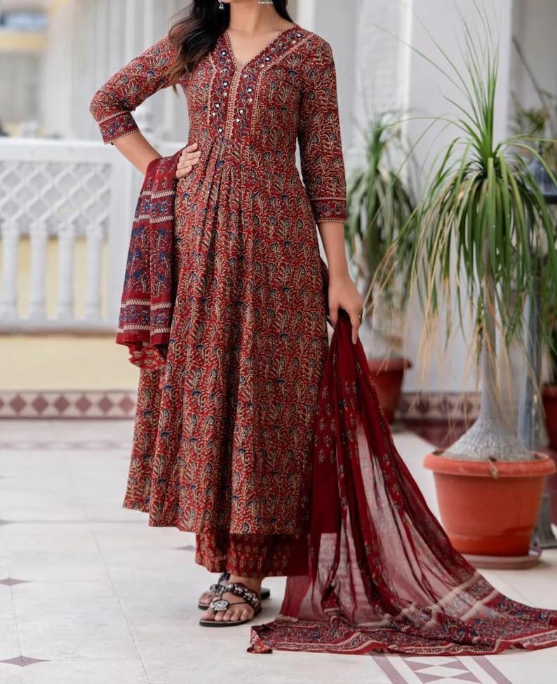 May include: A red floral print salwar kameez set. The long-sleeved top features a V-neck with decorative buttons and embroidery. The set includes matching pants and a sheer scarf. The outfit is worn with sandals.