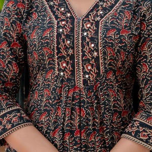 May include: Black and red floral print dress with a V-neckline. The dress features a detailed pattern with red flowers and leaves on a black background. The neckline and sleeves are trimmed with a beige border and small mirrors.