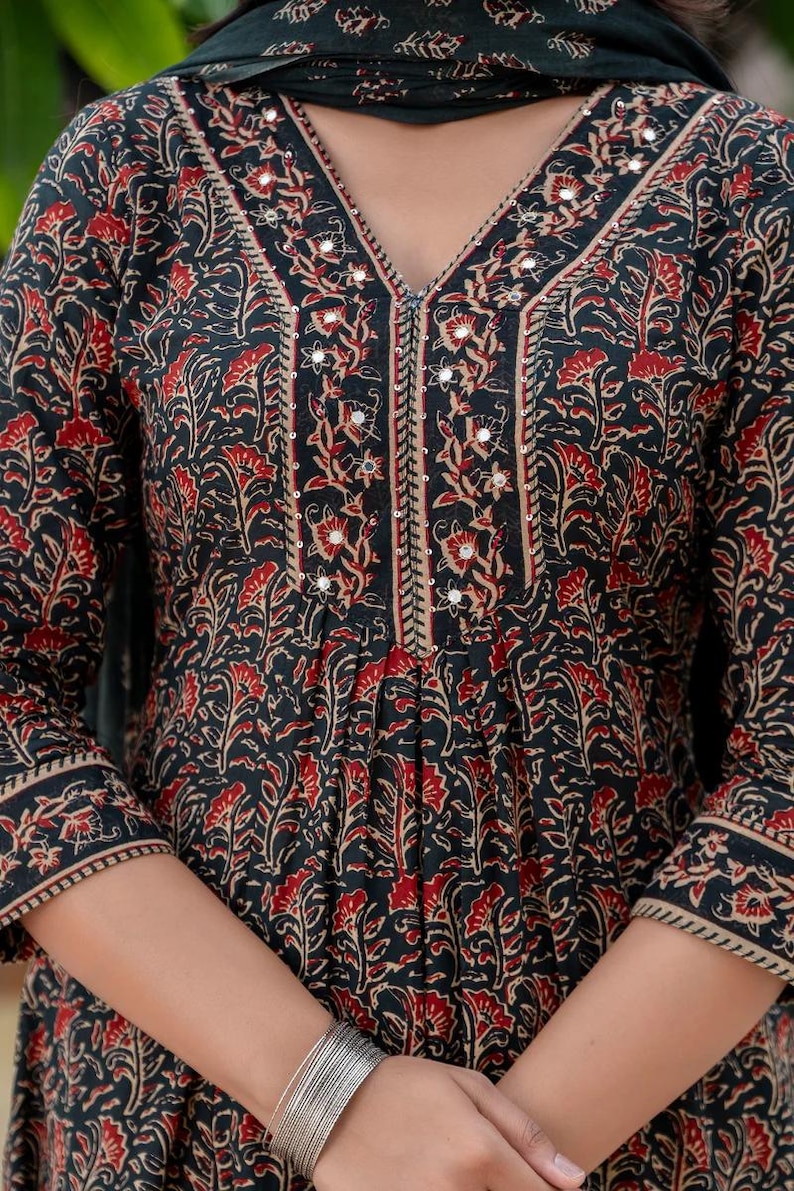 May include: Black and red floral print dress with a V-neckline. The dress features a detailed pattern with red flowers and leaves on a black background. The neckline and sleeves are trimmed with a beige border and small mirrors.