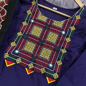 Puede incluir: Una t&uacute;nica morada con intrincados bordados en rojo, amarillo, verde y blanco. El dise&ntilde;o presenta patrones geom&eacute;tricos y peque&ntilde;os espejos. La t&uacute;nica se muestra en una percha de madera, mostrando la artesan&iacute;a detallada de la prenda.