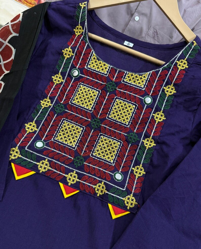 Puede incluir: Una t&uacute;nica morada con intrincados bordados en rojo, amarillo, verde y blanco. El dise&ntilde;o presenta patrones geom&eacute;tricos y peque&ntilde;os espejos. La t&uacute;nica se muestra en una percha de madera, mostrando la artesan&iacute;a detallada de la prenda.