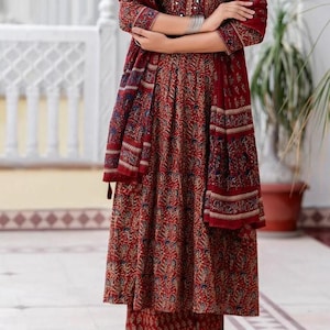 May include: A red floral print ensemble featuring a long-sleeved top, a flowing skirt, and a matching scarf. The outfit has a V-neckline and is paired with matching pants and sandals. The fabric has a detailed pattern with blue and beige accents.