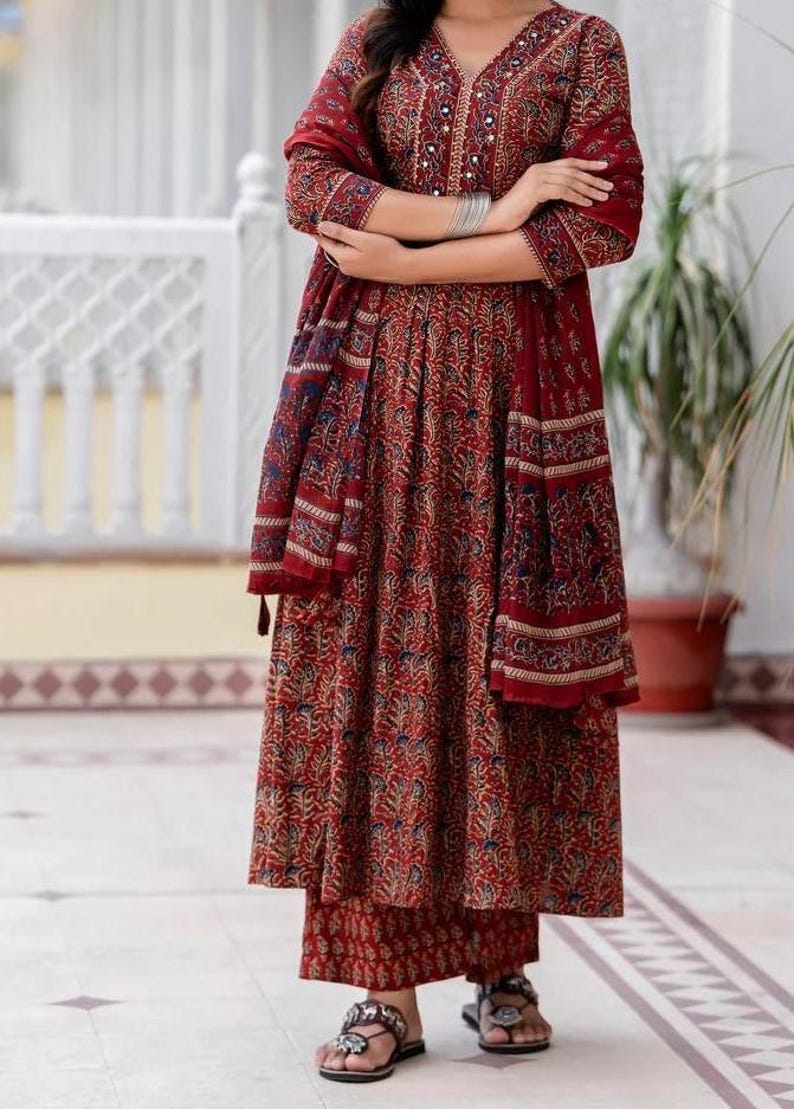 May include: A red floral print ensemble featuring a long-sleeved top, a flowing skirt, and a matching scarf. The outfit has a V-neckline and is paired with matching pants and sandals. The fabric has a detailed pattern with blue and beige accents.