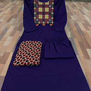 Puede incluir: Un conjunto morado de tres piezas, que incluye una t&uacute;nica de manga larga con detalles bordados en amarillo, rojo y verde, pantalones a juego y una bufanda estampada. La bufanda presenta un dise&ntilde;o geom&eacute;trico rojo, negro y blanco. El conjunto se exhibe sobre una superficie de madera.