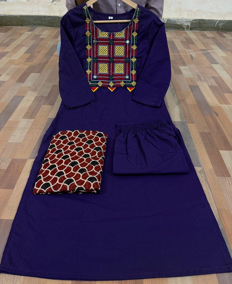 Puede incluir: Un conjunto morado de tres piezas, que incluye una t&uacute;nica de manga larga con detalles bordados en amarillo, rojo y verde, pantalones a juego y una bufanda estampada. La bufanda presenta un dise&ntilde;o geom&eacute;trico rojo, negro y blanco. El conjunto se exhibe sobre una superficie de madera.
