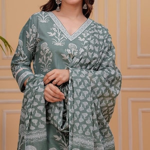 May include: A teal two-piece outfit with a matching scarf. The outfit features a V-neck top and long sleeves, both decorated with white floral patterns. The scarf and sleeves have a repeating tree-like design. The outfit is made of a soft, flowing fabric.