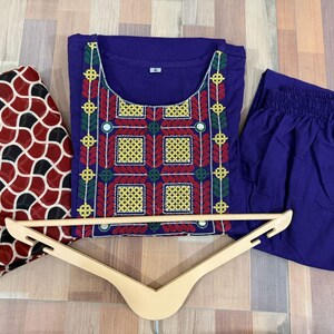 Puede incluir: Un conjunto de t&uacute;nica y pantal&oacute;n bordados en morado con una bufanda estampada. La t&uacute;nica presenta bordados coloridos en rojo, amarillo y verde. Una percha de madera se coloca delante de la ropa.