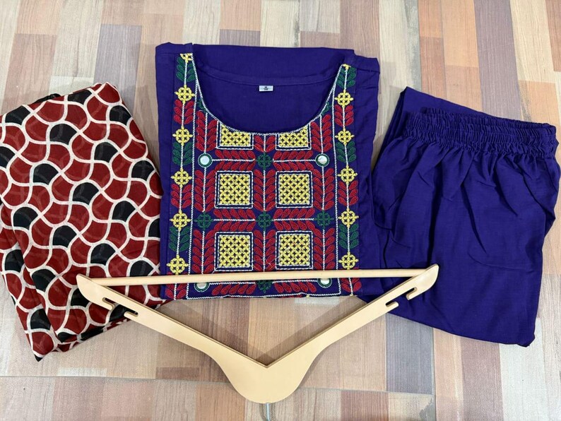 Puede incluir: Un conjunto de t&uacute;nica y pantal&oacute;n bordados en morado con una bufanda estampada. La t&uacute;nica presenta bordados coloridos en rojo, amarillo y verde. Una percha de madera se coloca delante de la ropa.