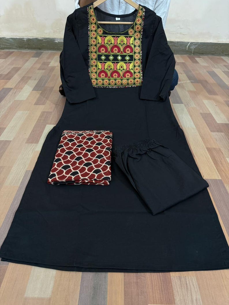 Puede incluir: Un conjunto de kurta negro con una parte superior de manga larga con un canes&uacute; bordado en rojo, amarillo y verde. El conjunto incluye pantalones negros a juego y una bufanda estampada en rojo y negro.