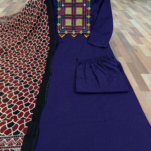 Puede incluir: Conjunto de t&uacute;nica y pantal&oacute;n morado oscuro con una bufanda estampada. La t&uacute;nica presenta bordados coloridos alrededor del escote. La bufanda tiene un patr&oacute;n repetido de formas rojas, negras y blancas.
