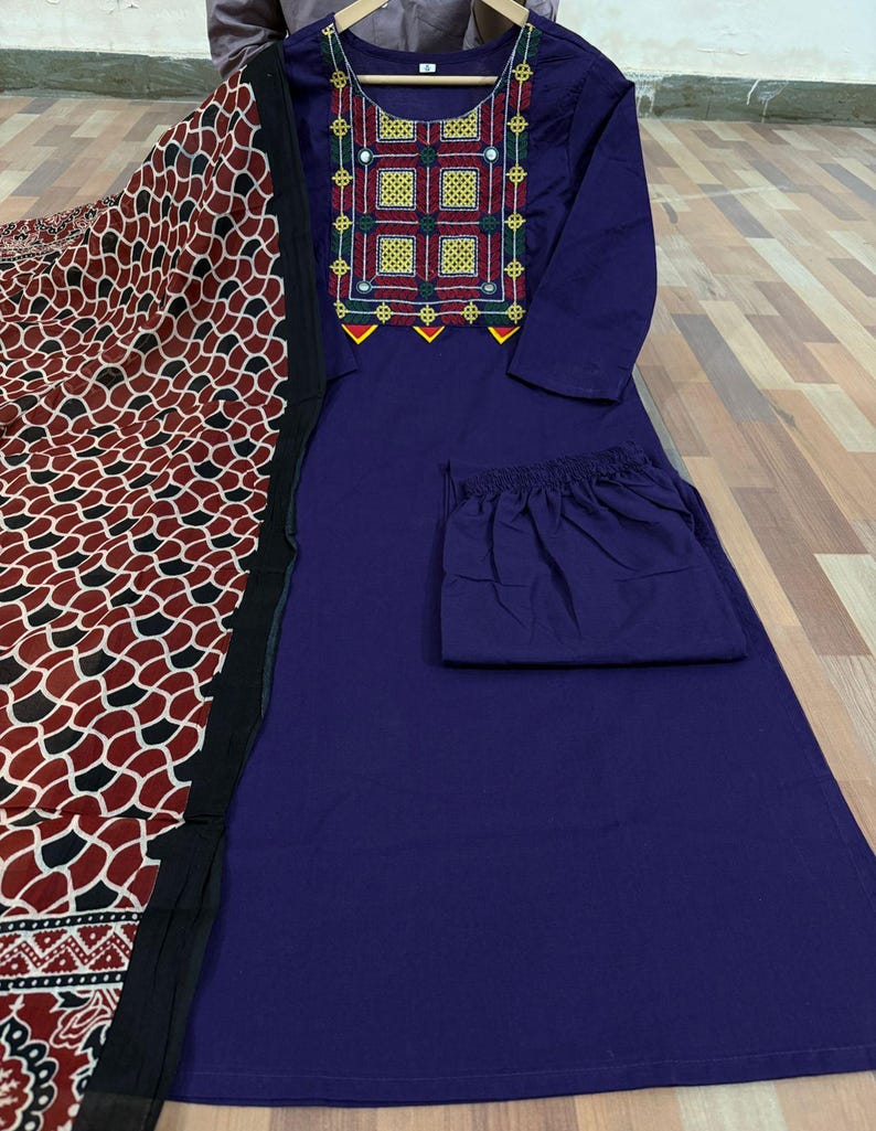 Puede incluir: Conjunto de t&uacute;nica y pantal&oacute;n morado oscuro con una bufanda estampada. La t&uacute;nica presenta bordados coloridos alrededor del escote. La bufanda tiene un patr&oacute;n repetido de formas rojas, negras y blancas.