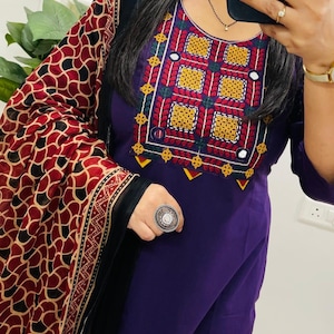 Puede incluir: Un salwar kameez morado con intrincados bordados en rojo, amarillo, verde y blanco alrededor del escote. El atuendo incluye un chal estampado con dise&ntilde;os rojos, negros y beige. Se ve un gran anillo plateado.