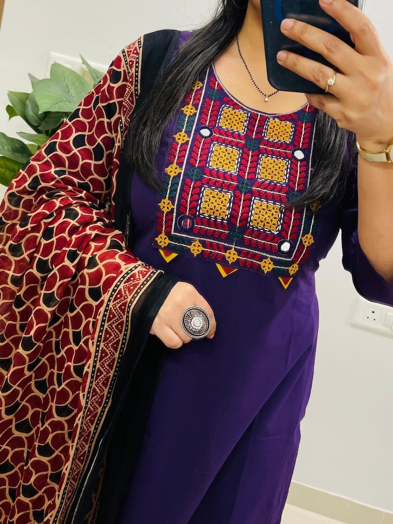 Puede incluir: Un salwar kameez morado con intrincados bordados en rojo, amarillo, verde y blanco alrededor del escote. El atuendo incluye un chal estampado con dise&ntilde;os rojos, negros y beige. Se ve un gran anillo plateado.
