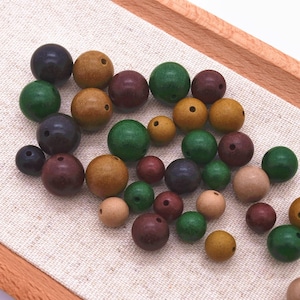 May include: A selection of wooden beads in various sizes and colours, including green, brown, tan, and purple. The beads are round with a hole drilled through the centre, and are scattered on a light-coloured fabric.