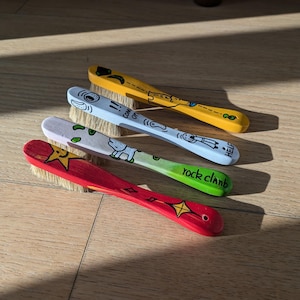 Puede incluir: Cuatro untadores de mantequilla pintados a mano en amarillo, azul claro, blanco y rojo. Cada untador tiene un diseño único y la frase "rock climb" en uno. Los untadores están sobre una superficie de madera. Miden unos 15 cm de largo.