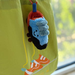 May include: A plush climbing shoe keychain in red, blue, and black hangs from a green tote bag with a carabiner clip. The bag features an orange climbing shoe graphic and the word "pelex" in white.